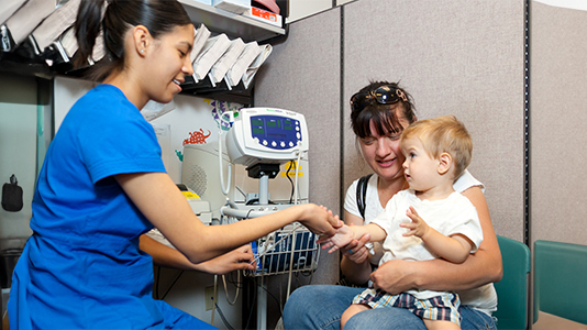 a healthcare worker examining a toddler's hand as the child sits on their mother's lap